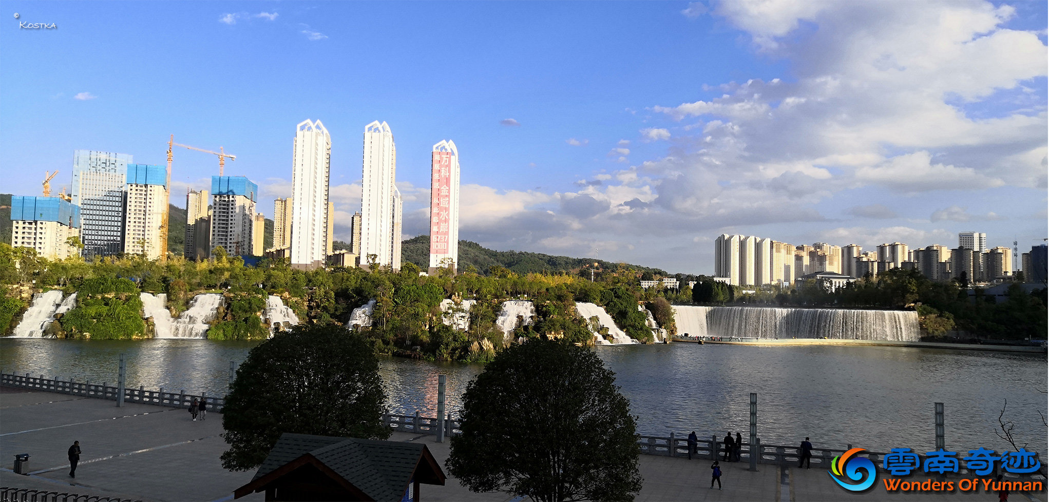 View from the west side platform over the lower lake of Waterfall Park with the 400-meter cascades and the natural waterfalls in the background people walking alongside the lake looking over the water watching the birds on the island 